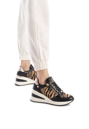 Zapatilla a rayas negras y marrones con suela gruesa, detalles metálicos y un cierre, combinada con pantalones blancos de puños elásticos.
