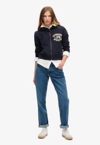 Marineblå zip-hoodie med "Superdry Japan Athletics" print, frontlomme, parret med opfoldede blå jeans og hvide sneakers.