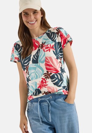 Kvinde smilende iført en beige kasket, en farverig t-shirt med tropisk bladprint og blå denim bukser med snøre, med den ene hånd i lommen.