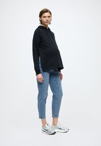 Μαύρο φούτερ με κουκούλα και πλάγιες σχισμές, φαρδιά εφαρμογή· ανοιχτό μπλε τζιν με φθορές· ασημένια και κρεμ αθλητικά παπούτσια με υφασμάτινο σχέδιο· λευκό φόντο.