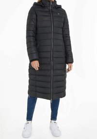 Long black puffer coat with a zip front, hood, and ribbed cuffs. Features horizontal quilted lines and a slim fit design. Worn with blue jeans and white sneakers.