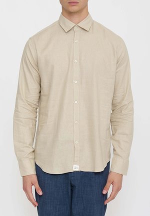 Uomo che indossa una camicia beige a maniche lunghe con colletto e pantaloni blu, in piedi davanti a uno sfondo semplice.