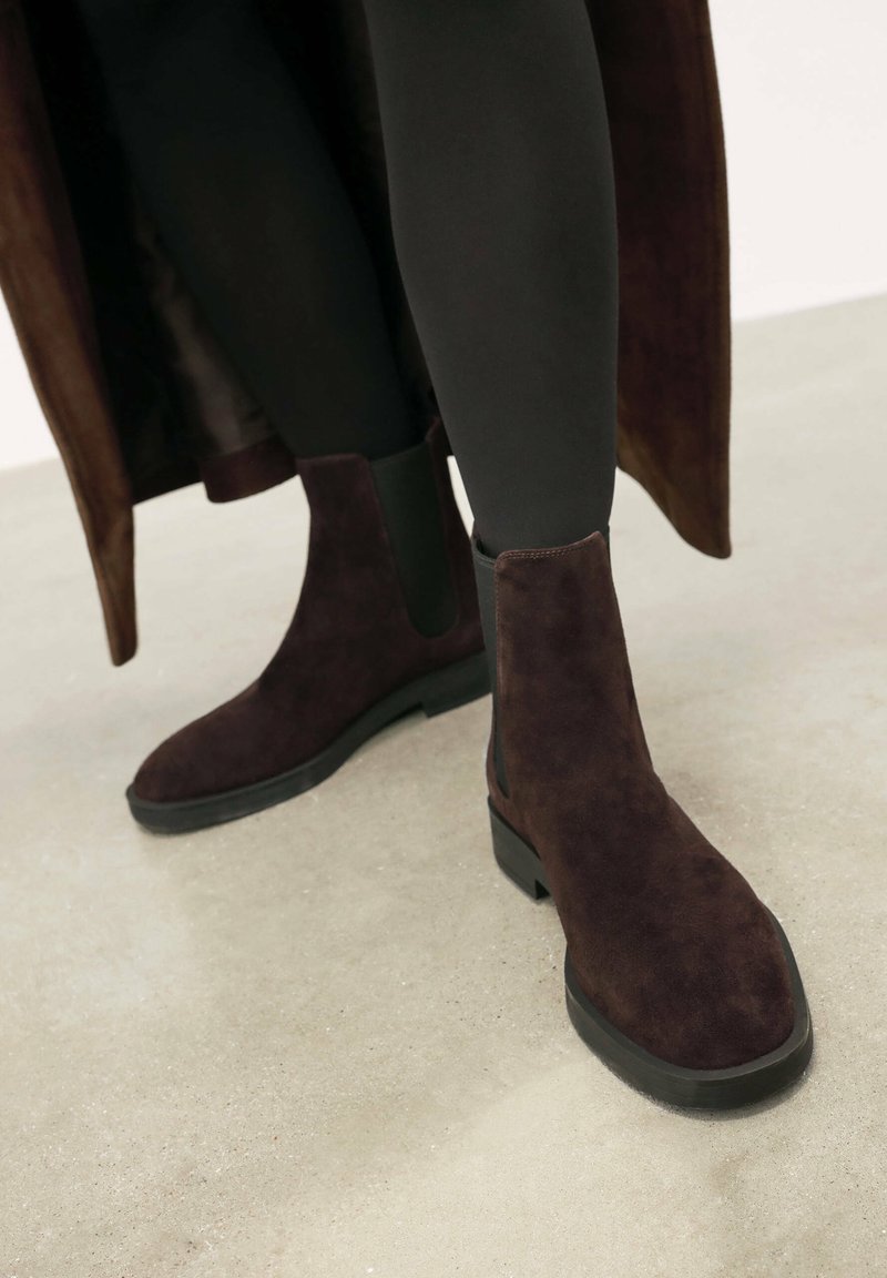 Boots din suede maro, la glezna, cu panouri laterale elastice, vârf rotund și toc mic pătrat, purtate cu colanți negri pe o suprafață neutră.