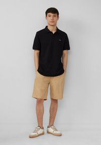 Schwarzes Poloshirt, khakifarbene Shorts und hellbraune Sneakers. Das Poloshirt hat einen Kragen und kurze Ärmel; die Shorts sind knielang mit seitlichen Taschen.