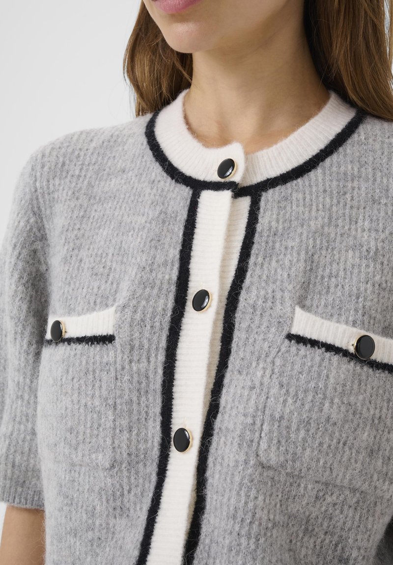 Cardigan gris en mélange de laine avec manches courtes, bordures noires et blanches, deux poches avant et boutons noirs. Tissu texturé offrant une sensation de confort.