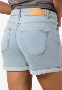 Pantalones cortos de denim azul claro con dobladillos enrollados, que cuentan con bolsillos traseros de parche y una etiqueta de cuero que dice "NOISY MAY" en la cintura.
