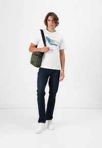 Hvit grafisk t-skjorte med blå og røde trykk, mørkeblå jeans og hvite joggesko. Holder en oliven grønn crossbody-veske.