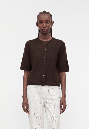 Femme aux cheveux tressés portant un pull à manches courtes boutonné marron foncé et un pantalon blanc avec des motifs découpés floraux.