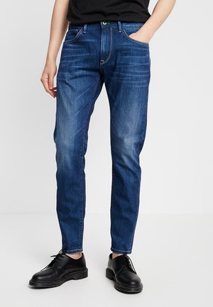 Person trägt dunkelblaue, eng anliegende Jeans und schwarze Schnürschuhe, steht mit einer Hand in der Tasche vor einem schlichten Hintergrund.