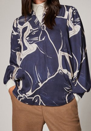 Kvinde iført en navyblå bluse med store abstrakte katteprint, hvid rullekrave under, og brune bukser med hænderne i lommerne.
