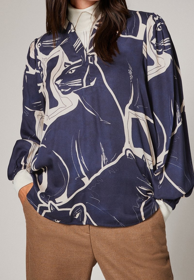 Femme portant un chemisier bleu marine avec de grands imprimés abstraits de chats, un col roulé blanc en dessous, et un pantalon marron avec les mains dans les poches.