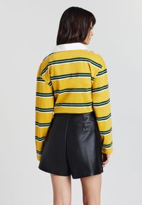 Frau, die von hinten schaut, trägt ein langärmliges Hemd mit gelben, weißen und grünen Streifen und einem weißen Kragen sowie schwarze Leder-Shorts.