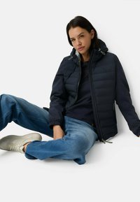 Marineblauwe pufferjack met capuchon, ritssluiting en elastische manchetten. Combinatie met lichtblauwe spijkerbroek en witte sneakers, zittend op een effen achtergrond.