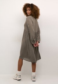 Vestito khaki con pois bianchi, maniche lunghe e silhouette fluida. Il modello indossa sneakers e calze con risvolto abbassato.