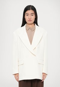 Dvojradové biele sako z pevnej látky s širokými klopami, šikmými vreckami a voľným strihom. Nosí sa cez béžovú košeľu s golierom.