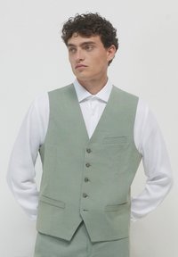 Hellgrüne Weste aus strukturiertem Stoff, fünf Knöpfe und zwei Taschen vorne, kombiniert mit einem weißen Hemd mit Kragen. Glatter Halsausschnitt und taillierte Passform.