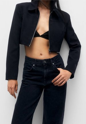 Frau trägt eine kurze schwarze Reißverschlussjacke, ein schwarzes Bustier und hoch taillierte schwarze Jeans, mit einer Hand in der Tasche vor weißem Hintergrund.