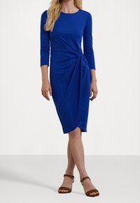 Knälång blå klänning med lång ärm, med en snodd detaljerad framsida och mjuk tygtextur. Matchad med bruna sandaler med remmar.