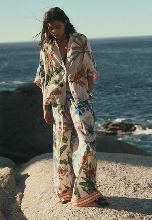 Nő áll egy sziklás parton, laza virágmintás ingben és bő szárú nadrágban, háttérben az óceán.