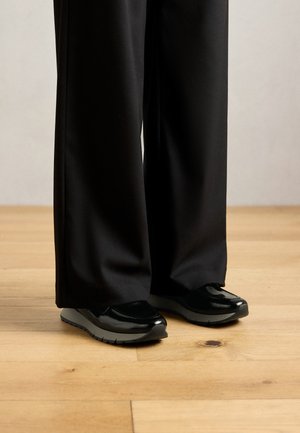 Schwarze weit geschnittene Hosen, die die Knöchel bedecken, kombiniert mit glänzenden schwarzen Sneakers auf einem hellen Holzboden.