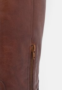 Bota tobillera de cuero marrón con un acabado texturizado, que presenta un cierre lateral, costuras sutiles y un diseño de punta redondeada.