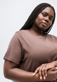 Νεαρή μαύρη γυναίκα με μακριά πλεξούδες, που φοράει ένα καφέ κοντομάνικο πουκάμισο, τα χέρια της απαλά σφιγμένα, κοιτάζει με αυτοπεποίθηση την κάμερα πάνω σε απλό φόντο.