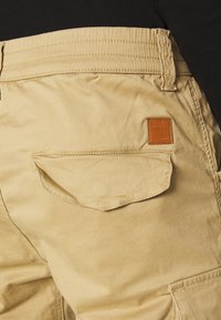 Beige cargobyxor med en bakficka med rund flap, sydda detaljer och en brun läderlogotyp på midjebandet.