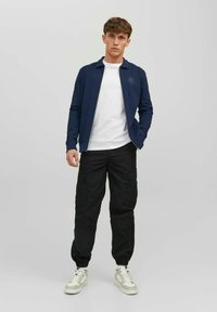 Veste zippée marine avec col, associée à un T-shirt blanc, un pantalon cargo noir et des baskets blanches, offrant une coupe décontractée et des détails minimalistes.