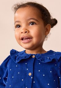 Robe en velours côtelé bleu à pois blancs, avec un col à volants, une fermeture à boutons et un tissu texturé. Le modèle sourit devant un fond neutre.