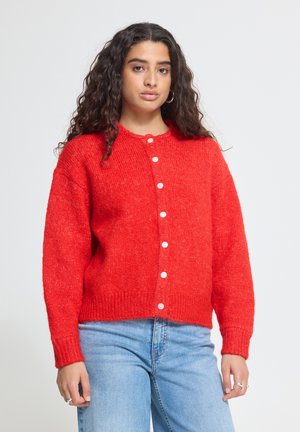 Kvinde med langt krøllet hår iført en knap-foret, knaldrød cardigan og lyseblå jeans, stående foran en ensfarvet baggrund.