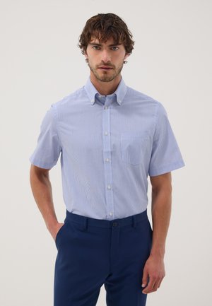 Man met krullend bruin haar die een lichtblauw kortärmelig overhemd en donkerblauwe broek draagt, staand tegen een effen achtergrond.