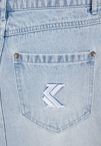 Tissu en denim bleu clair avec une finition texturée. Comprend une poche arrière avec des accessoires en métal argenté et un détail de logo brodé en blanc.