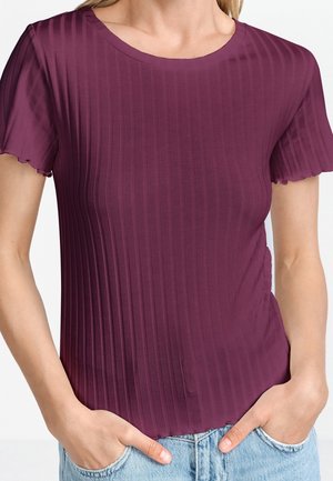 Kobieta ubrana w ciemnofioletowy, prążkowany top z krótkim rękawem i okrągłym dekoltem, włożony do jasnoniebieskich dżinsów, ręce w kieszeniach.