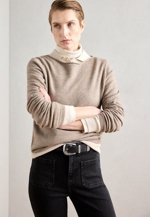 Beżowy wełniany sweter nałożony na kremową koszulę polo, w połączeniu z czarnymi dżinsami. Zawiera czarny pasek z srebrną klamrą. Miękka tekstura, luźny krój.