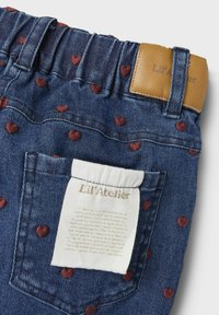 Denim trousers featuring a soft elastic waistband, brown leather tag, and maroon heart patterns on a blue background. Care label visible on pocket.