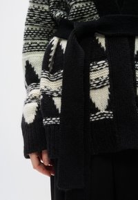 Πλεκτή ζακέτα ασπρόμαυρη με γεωμετρικά σχέδια, ζωνάκι δεμένο στη μέση και υφή υφάσματος που φαίνεται ζεστή και παχιά.