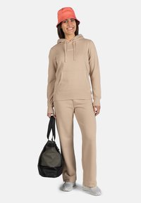 Sudadera beige y pantalones de chándal a juego hechos de tela suave. La persona sostiene un bolso de deporte negro y gris con dos correas. Un sombrero de cubo coral completa el look.