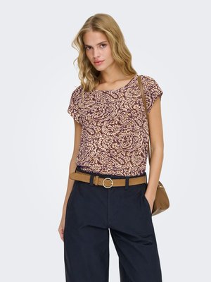 Blonde woman wearing a patterned short-sleeve top, navy pants with tan belt, and a tan shoulder bag, standing against plain background.