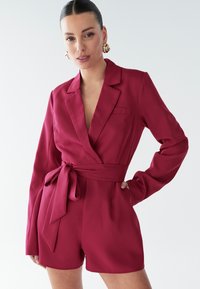 Fľaškovozelený zavinovací blejzerový overal vyrobený z hladkej látky. Má hlboký výstrih do V, dlhé rukávy, opasok na viazanie a bočné vrecká.