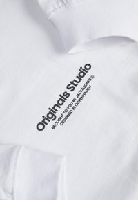 Vit bomull t-shirt med svart tryckt text "Originals Studio Brought to you by Jack & Jones Designad i Köpenhamn." Långa ärmar.