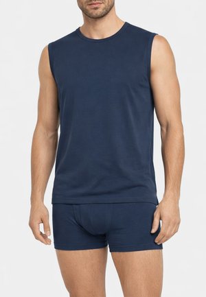 Uomo che indossa una canottiera blu navy senza maniche e boxer aderenti abbinati, in piedi contro uno sfondo chiaro e uniforme.