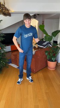 Eine Person trägt ein blaues T-Shirt mit einem weißen Motiv, blaue Jeans und marineblaue Sneaker. Sie hält ein schwarzes Kleidungsstück mit weißem Besatz.