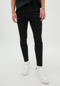 Jeans ajustados negros, de tela de mezcla de algodón, con diseño de cinco bolsillos y una textura suave. Combinados con zapatillas blancas con suelas texturizadas.