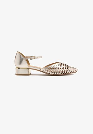 Chaussure plate en métal doré avec un bout pointu, un design découpé, une sangle à la cheville et un petit talon carré. Fini lisse et brillant.