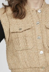 Beige tekstureret vest med to forlommer og sølvknapper. Materialet har et vævet mønster, der skaber et struktureret look.