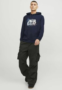 Sudadera azul marino con un estampado gráfico blanco y cordones, combinada con pantalones cargo negros que cuentan con bolsillos laterales y un ajuste holgado.