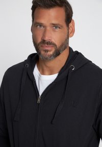 Sudadera negra con cremallera y cordones, que presenta un logotipo sutil en el pecho. Superpuesta a una camiseta blanca, hecha de un tejido suave.