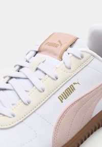 Baskets blanches avec des accents en daim rose et crème, logo doré et lacets clairs. Présentent une semelle en caoutchouc texturée et une languette rembourrée.