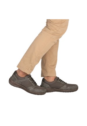 Person mit beigen Hosen und grauen lässigen Schnürschuhen, zeigt überkreuzte Beine in der Mitte eines Schritts vor weißem Hintergrund.