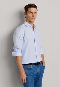 Mężczyzna w jasno-niebieskiej koszuli w prążki z podwiniętymi rękawami i niebieskich dżinsach, stojący z rękami w kieszeniach na tle szarej ściany.